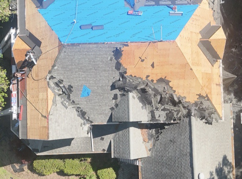 Before: aerial view of a large home stripped to bare plywood decking, with torn-off shingles piled on the lower slopes and blue synthetic underlayment across the upper roof